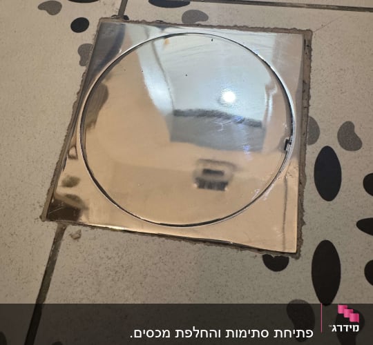 פתח ניקוז עגול ברצפת אריחים עם כיסוי מתכת מבריק.